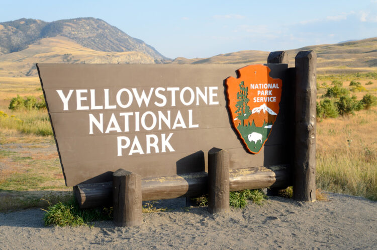 Alt text: "Welcome sign for Yellowstone National Park featuring bold, white lettering on a dark background, with the National Park Service emblem to the right side, set against a backdrop of rolling hills and a clear blue sky."
