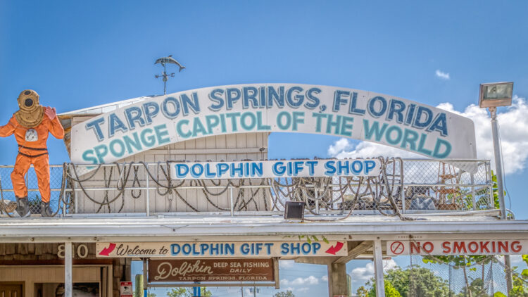 Alt text: "Photograph of the iconic Tarpon Springs Sponge Docks sign reading 'Tarpon Springs, Florida - Sponge Capital of the World' with a life-size diver statue and the Dolphin Gift Shop entrance below. Welcoming visitors to explore deep