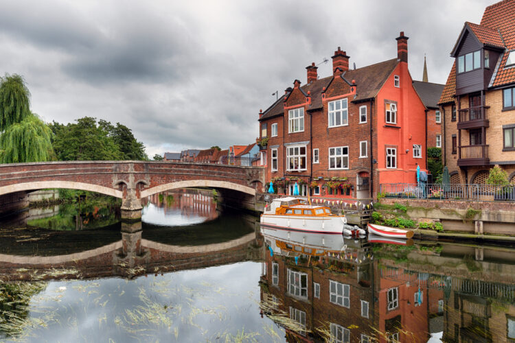 "Quaint European town with historic red brick architecture and charming canal boats reflected in the calm water under an arched stone bridge, evoking a sense of peaceful exploration for travelers."