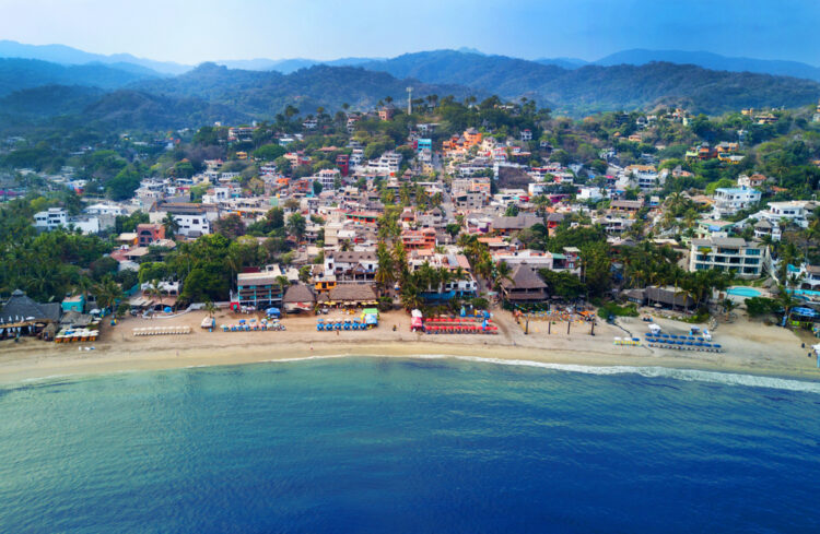 Alt text: "Aerial view of a picturesque coastal village with dense housing flanked by lush green hills, facing a tranquil blue sea with a sandy beach lined by colorful umbrellas and palm trees, embodying a perfect tropical paradise for travelers seeking serene