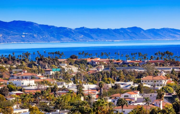 "Panoramic view of a coastal town with palm trees, distinctive red-tiled roofs, and lush greenery against a backdrop of serene blue ocean and distant mountains under clear skies, perfect for a tranquil getaway." Please note that this alt text