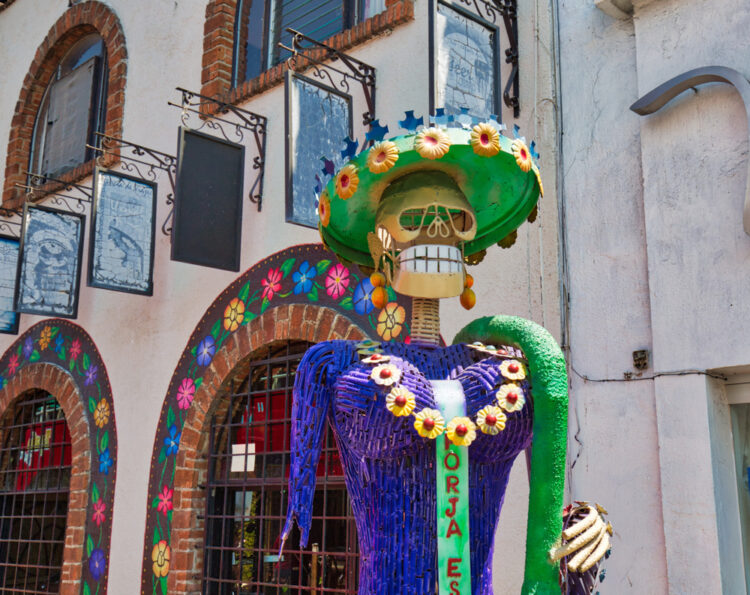"Colorful Catrina sculpture with flower-adorned hat in a vibrant Mexican street setting, exemplifying traditional Day of the Dead art and culture for an immersive travel experience."