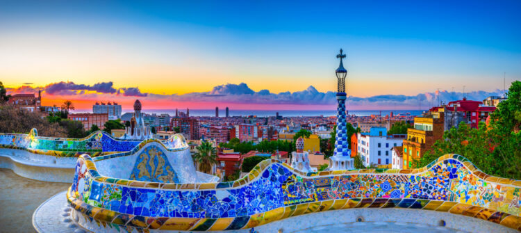 "Panoramic view at sunset of the vibrant mosaic bench at Park Güell with Barcelona cityscape and coastline in the background, showcasing Gaudi's modernist architecture and breathtaking Spanish urban vistas."